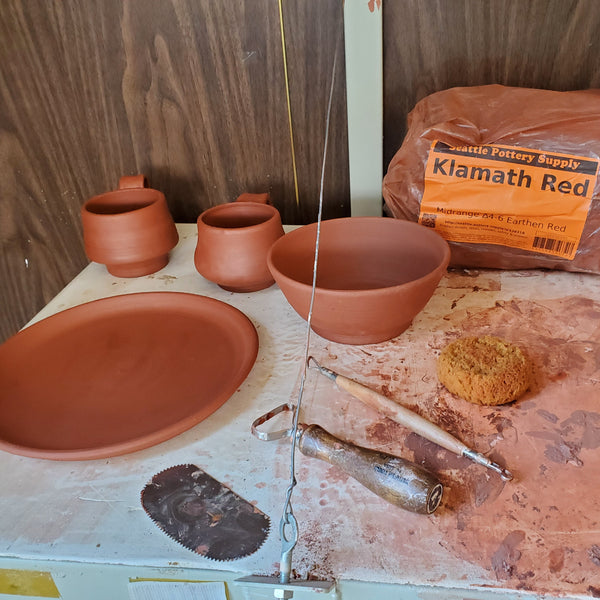 A bowl,  plate, and two mugs made of Happycozygoods Klamath Red Pottery clay sit on a shelf with pottery tools