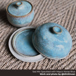 Two lidded bowls made from Happycozygoods sea mix speckled clay and norse blue glaze on a woven surface